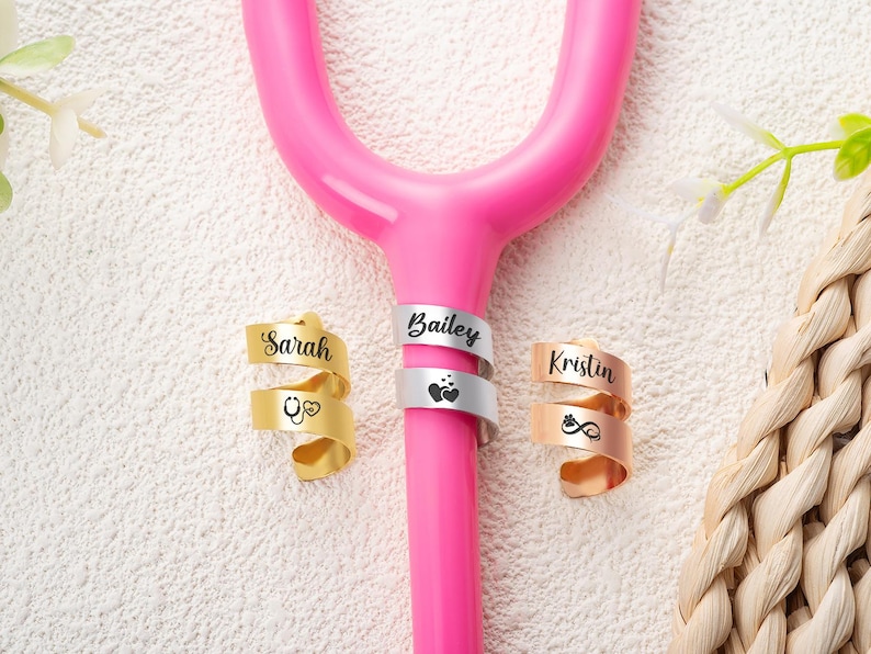 May include: A bright pink stethoscope tubing section adorned with three spiral metal identification tags. The gold tag is engraved with Sarah and a stethoscope icon, the silver tag with Bailey and hearts, and the rose gold tag with Kristin and a paw print.