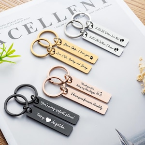 May include: Four pairs of rectangular metal keychains in silver, gold, rose gold, and black finishes. Each keychain features a split ring and a flat bar engraved with custom text, including dates, names, and phrases like 'Drive Safe' and 'I love you'.