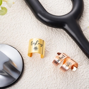 May include: Two metal cuff rings with open bands. One gold-toned ring is engraved with the name Lesley and a sun icon. One rose gold-toned ring is engraved with the name Rachel and a stethoscope icon. The rings are positioned near a stethoscope.