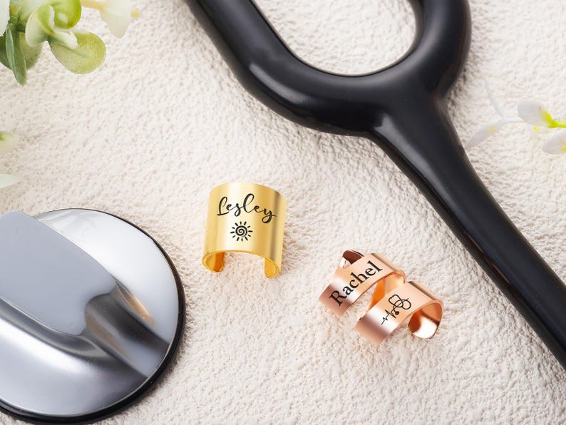 May include: Two metal cuff rings with open bands. One gold-toned ring is engraved with the name Lesley and a sun icon. One rose gold-toned ring is engraved with the name Rachel and a stethoscope icon. The rings are positioned near a stethoscope.