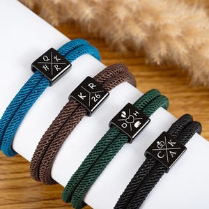 Op de afbeelding: Vier gevlochten touwarmbanden in teal, bruin, groen en zwart. Elke armband heeft een zwarte vierkante sluiting met witte gegraveerde ontwerpen, waaronder initialen, cijfers en symbolen. De armbanden worden op een wit oppervlak weergegeven.