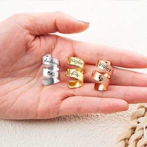 May include: Three spiral-shaped metal rings in silver, gold, and rose gold tones. Each ring features a custom engraving: the silver ring displays the name Andrew and an infinity symbol, the gold ring shows Naylor and a heartbeat, and the rose gold ring shows Violet and baby footprints.