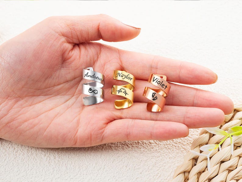 May include: Three spiral-shaped metal rings in silver, gold, and rose gold tones. Each ring features a custom engraving: the silver ring displays the name Andrew and an infinity symbol, the gold ring shows Naylor and a heartbeat, and the rose gold ring shows Violet and baby footprints.
