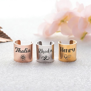 May include: Three open-band rings in rose gold, silver, and gold tones. Each ring features a custom name engraving: Thalia with a sun icon, Rhoda with an infinity and paw print symbol, and Henry with a stethoscope and heart icon in black ink.