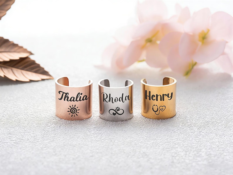 May include: Three open-band rings in rose gold, silver, and gold tones. Each ring features a custom name engraving: Thalia with a sun icon, Rhoda with an infinity and paw print symbol, and Henry with a stethoscope and heart icon in black ink.