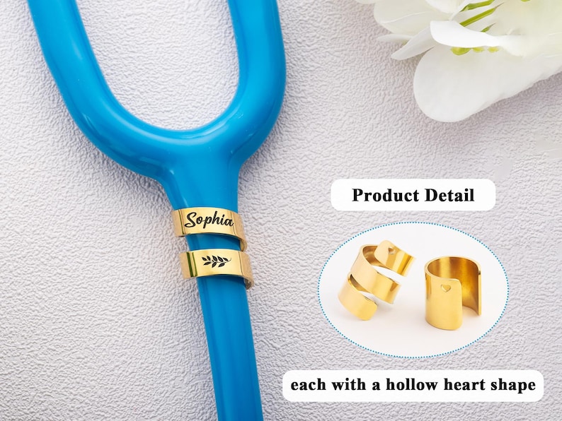 May include: A gold-coloured metal identification tag for a stethoscope, featuring the name Sophia and a leaf engraving. The product detail inset shows two gold-coloured metal tags, each with a small hollow heart shape cutout on the side.