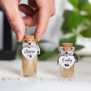 May include: Two small glass vials with cork stoppers, filled with light brown material. Each vial has a heart-shaped tag with a name and paw print. One says "Oliver" and the other says "Emily". The vials are displayed on a white, textured surface.