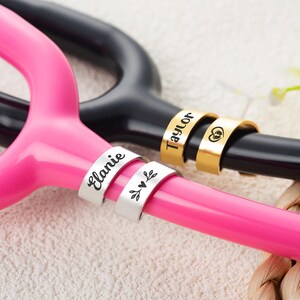 May include: Two metal identification tags attached to a bright pink stethoscope and two gold-toned tags on a black stethoscope. The silver tags are engraved with the name Elanie and a leaf design. The gold tags feature the name Taylor and a heart icon.