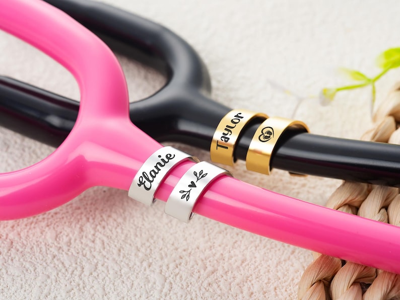May include: Two metal identification tags attached to a bright pink stethoscope and two gold-toned tags on a black stethoscope. The silver tags are engraved with the name Elanie and a leaf design. The gold tags feature the name Taylor and a heart icon.