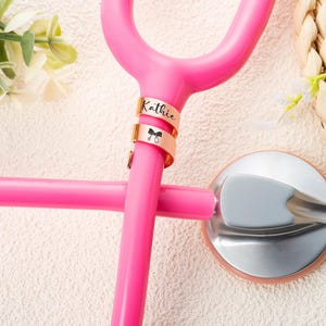 May include: A bright pink stethoscope featuring two rose gold metal identification tags attached to the tubing. The top tag is engraved with the name Kathie, and the bottom tag displays a small black icon of a stethoscope with a bow.
