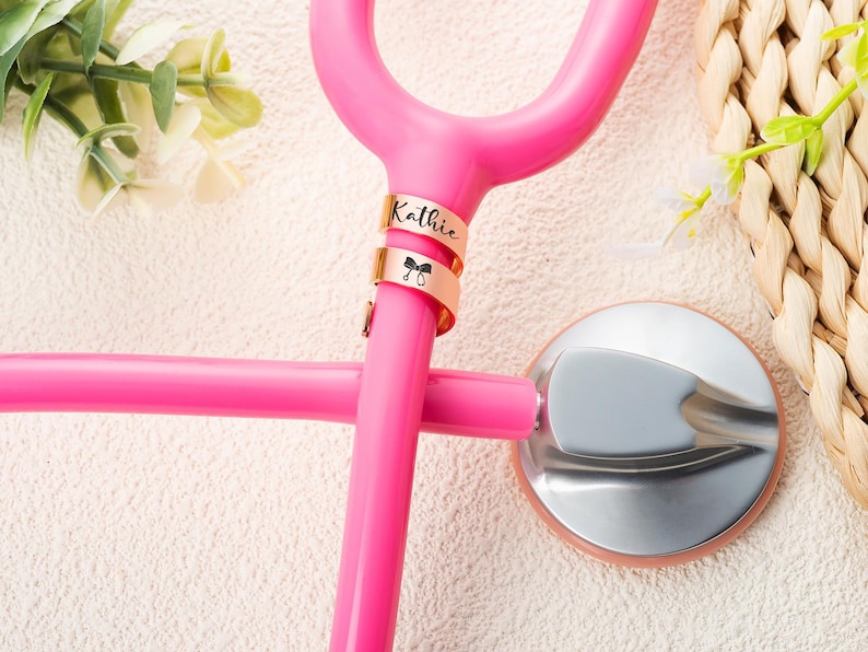 May include: A bright pink stethoscope featuring two rose gold metal identification tags attached to the tubing. The top tag is engraved with the name Kathie, and the bottom tag displays a small black icon of a stethoscope with a bow.