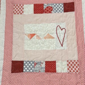 May include: A handmade quilt with a heart design. The quilt features a red and white polka dot border and a central square with a heart outline. The square has a light pink background with a heart applique and geometric shapes.