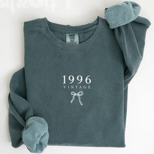 May include: A blue spruce sweatshirt with the year "1996" and the word "VINTAGE" printed in white. A white bow graphic is below the text. The sleeves are cuffed, and the sweatshirt is folded. The words "Dal&Gift" and "Blue Spruce" are also visible.