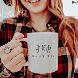 May include: White ceramic mug with a black skeleton design and the word "RADIOLOGY". The mug also has the text "CERAMIC MUG" and "11oz" printed on it. The mug is held by a person wearing a plaid shirt.