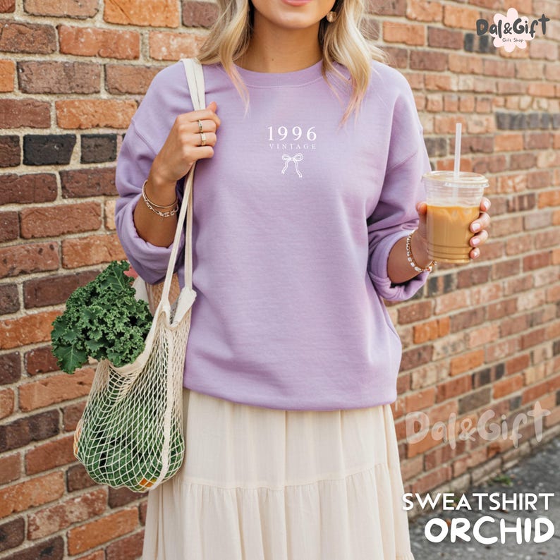 May include: A lilac-colored sweatshirt with the text "1996 Vintage" and a bow graphic. The sweatshirt is part of a casual outfit, paired with a cream-colored skirt. The image also shows a tote bag with produce and a drink.