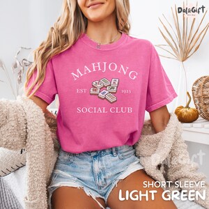 May include: A pink short-sleeve t-shirt with the text "MAHJONG SOCIAL CLUB" and a graphic of mahjong tiles. The shirt also has the text "EST 1923". The shirt is made of a soft material.