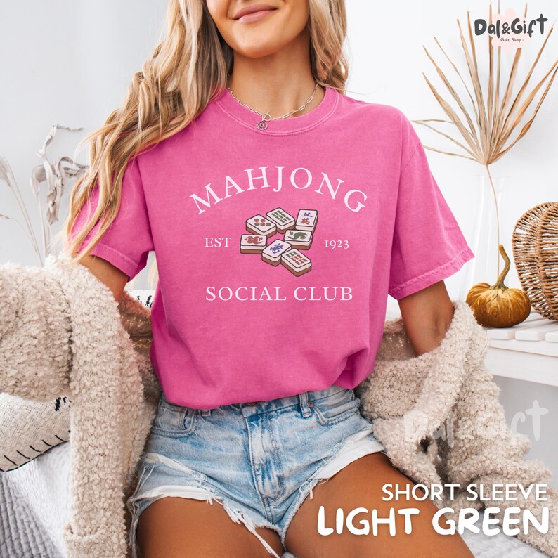 May include: A pink short-sleeve t-shirt with the text "MAHJONG SOCIAL CLUB" and a graphic of mahjong tiles. The shirt also has the text "EST 1923". The shirt is made of a soft material.