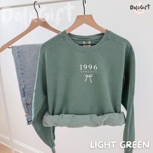 May include: A light green sweatshirt with the text "1996 VINTAGE" and a bow graphic. The sweatshirt is displayed on a wooden hanger next to a pair of blue jeans. The words "LIGHT GREEN" are at the bottom of the image.