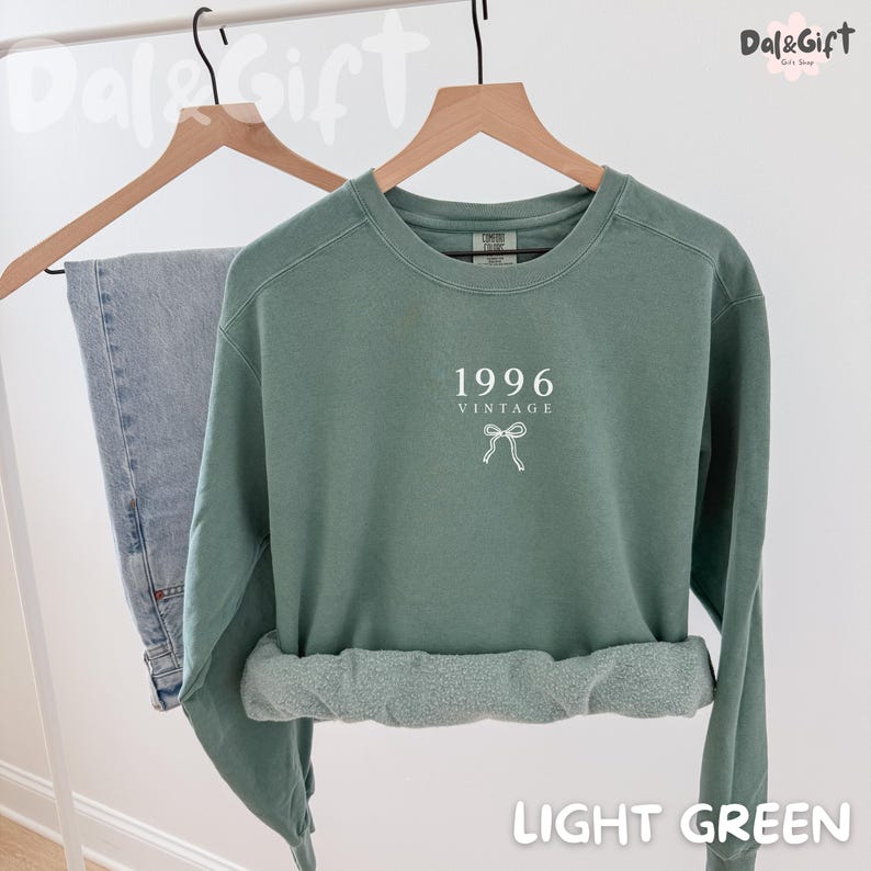 May include: A light green sweatshirt with the text "1996 VINTAGE" and a bow graphic. The sweatshirt is displayed on a wooden hanger next to a pair of blue jeans. The words "LIGHT GREEN" are at the bottom of the image.