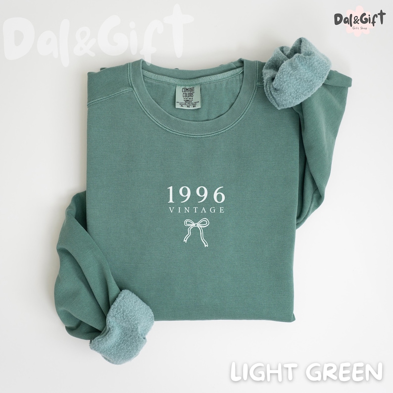 May include: Light green sweatshirt with the year "1996" and the word "VINTAGE" printed in white. A white bow graphic is below the text. The sweatshirt is folded, with the sleeves showing a soft, textured lining.