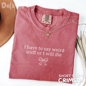 May include: Crimson short-sleeve t-shirt with the text "I have to say weird stuff or I will die" and a bow graphic. A silver necklace with a pendant is draped on the shirt. The shirt is made by Comfort Colors.