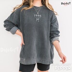May include: A dark gray vintage-style sweatshirt with the year "1996" and a bow graphic printed on the front. The sweatshirt has a crew neck and long sleeves with rolled-up cuffs. The word "SWEATSHIRT" and the color "PEPPER" are printed on the bottom right.