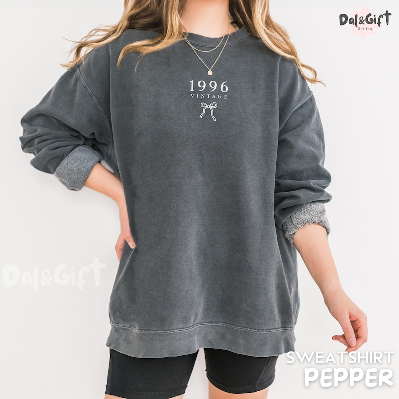 May include: A dark gray vintage-style sweatshirt with the year "1996" and a bow graphic printed on the front. The sweatshirt has a crew neck and long sleeves with rolled-up cuffs. The word "SWEATSHIRT" and the color "PEPPER" are printed on the bottom right.