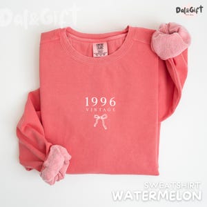 May include: A watermelon-colored sweatshirt with the text "1996 VINTAGE" and a bow graphic. The sweatshirt has a crew neck and long sleeves with a soft, textured cuff. The brand name "Comfort Colors" is visible on the tag.