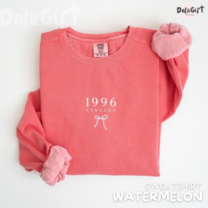 May include: A watermelon-colored sweatshirt with the text "1996 VINTAGE" and a bow graphic. The sweatshirt has a crew neck and long sleeves with a soft, textured cuff. The brand name "Comfort Colors" is visible on the tag.