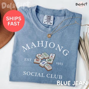 May include: A blue jean colored t-shirt with the words "MAHJONG SOCIAL CLUB" printed on the front. The shirt features a graphic of Mahjong tiles and the text "EST 1923". A gold necklace with a pendant is draped across the shirt.