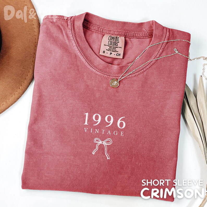 May include: Crimson short-sleeve t-shirt with the text "1996 VINTAGE" and a bow graphic. The shirt has a crew neck and a Comfort Colors tag. A silver necklace with a pendant is draped across the shirt.