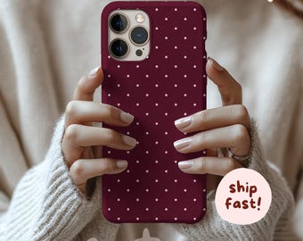 May include: A burgundy phone case with a white polka dot pattern. The case is held by a person wearing a cream-colored sweater. The case has a camera cutout and the words "ship fast!" on a sticker.