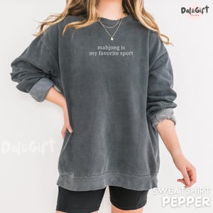 Pu&ograve; includere: Una felpa grigio scuro con la scritta bianca "mahjong is my favorite sport". La felpa ha maniche lunghe con polsini arrotolati. La parola "SWEATSHIRT" e il colore "PEPPER" sono stampati in basso.