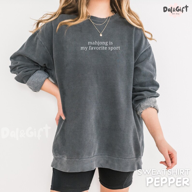 Pu&ograve; includere: Una felpa grigio scuro con la scritta bianca "mahjong is my favorite sport". La felpa ha maniche lunghe con polsini arrotolati. La parola "SWEATSHIRT" e il colore "PEPPER" sono stampati in basso.