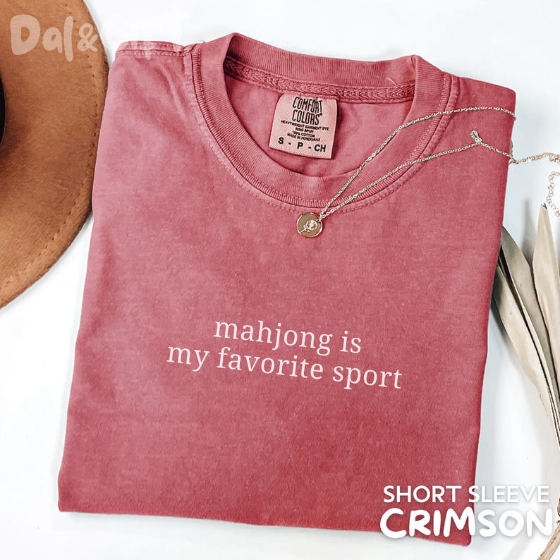 Pu&ograve; includere: T-shirt a maniche corte color cremisi con la scritta bianca "mahjong is my favorite sport". Una collana d'argento con un ciondolo &egrave; drappeggiata sulla maglietta. La maglietta ha un'etichetta Comfort Colors.