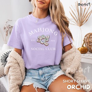 May include: A light purple short-sleeve t-shirt with the words "MAHJONG SOCIAL CLUB" and a graphic of Mahjong tiles. The text "EST 1923" is also included. The shirt is a soft, comfortable material.