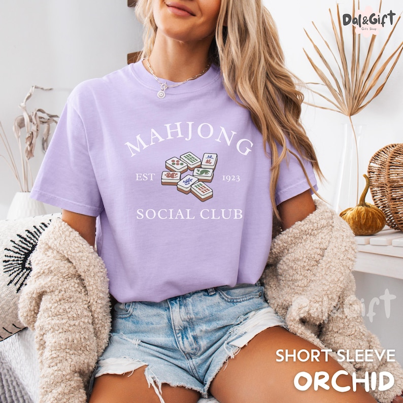 May include: A light purple short-sleeve t-shirt with the words "MAHJONG SOCIAL CLUB" and a graphic of Mahjong tiles. The text "EST 1923" is also included. The shirt is a soft, comfortable material.