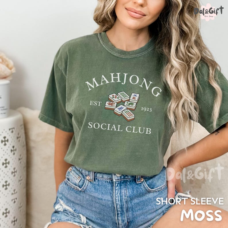 May include: Moss green short-sleeve t-shirt with the text "MAHJONG SOCIAL CLUB EST 1923" in white. The shirt features a graphic of mahjong tiles. The shirt is made of a soft material.