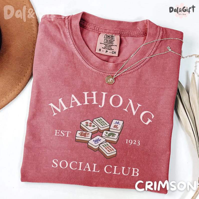 May include: Crimson-colored t-shirt with the text "MAHJONG SOCIAL CLUB EST 1923" and a graphic of Mahjong tiles. The shirt has a crew neck and short sleeves. A gold necklace with a pendant is draped on the shirt.