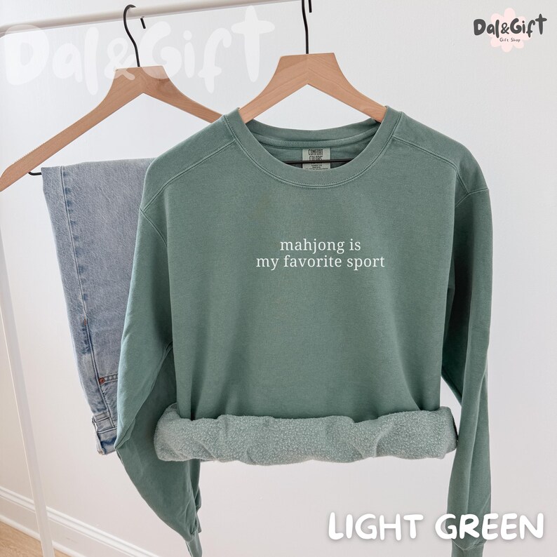Pu&ograve; includere: Felpa verde chiaro con la scritta bianca "mahjong is my favorite sport". La felpa &egrave; appesa a una gruccia di legno accanto a un paio di jeans blu. Il fondo della felpa &egrave; arrotolato.