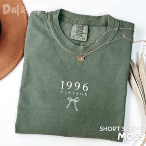 May include: A moss green short-sleeve t-shirt with the text "1996 VINTAGE" and a bow graphic. The shirt has a crew neck and a small tag on the upper back. A gold necklace with a pendant is draped across the shirt.