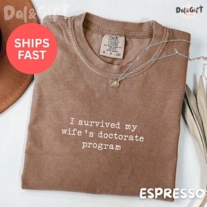 May include: Espresso-colored t-shirt with the text "I survived my wife's doctorate program" in white. The shirt has a crew neck and short sleeves. A silver necklace with a pendant is draped across the shirt.