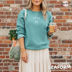 May include: Seafoam green sweatshirt with the text "1996 Vintage" and a bow graphic. The sweatshirt is paired with a cream-colored skirt. A reusable shopping bag filled with produce is visible. The image also shows a drink in a clear cup.