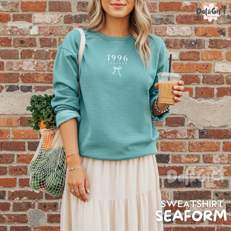 May include: Seafoam green sweatshirt with the text "1996 Vintage" and a bow graphic. The sweatshirt is paired with a cream-colored skirt. A reusable shopping bag filled with produce is visible. The image also shows a drink in a clear cup.