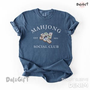 May include: A denim blue short-sleeve t-shirt with the words "MAHJONG SOCIAL CLUB EST 1923" printed in white. The graphic includes several Mahjong tiles. The shirt is a casual style.
