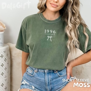 May include: Olive green short-sleeve t-shirt with the text "1996 VINTAGE" and a bow graphic. The shirt is made of a soft material and is paired with denim shorts. The bottom right corner of the shirt reads "SHORT SLEEVE MOSS".