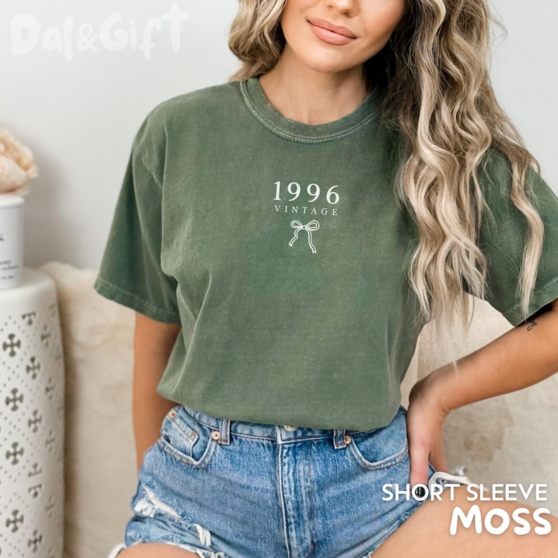 May include: Olive green short-sleeve t-shirt with the text "1996 VINTAGE" and a bow graphic. The shirt is made of a soft material and is paired with denim shorts. The bottom right corner of the shirt reads "SHORT SLEEVE MOSS".