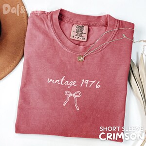 May include: Crimson short-sleeve t-shirt with the text "vintage 1976" and a bow graphic. A silver necklace with a pendant is draped on the shirt. The shirt has a Comfort Colors tag.