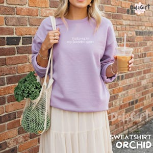 Pu&ograve; includere: Una felpa lilla con la scritta bianca "mahjong is my favorite sport". La felpa fa parte della collezione "Sweatshirt Orchid". La persona nell'immagine tiene in mano una borsa della spesa riutilizzabile con prodotti e una bevanda al caff&egrave;.