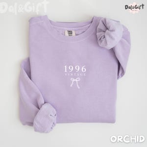 May include: A light purple sweatshirt with the text "1996 VINTAGE" and a bow graphic. The sweatshirt is a solid color and has a crew neck. The sleeves are cuffed. The word "ORCHID" is at the bottom right.